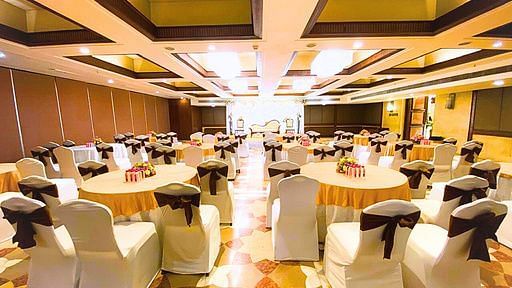 A well-lit banquet hall at Pride Plaza Ahmedabad, featuring a large number of round tables with white tablecloths and black chair covers, set up for a conference or reception.